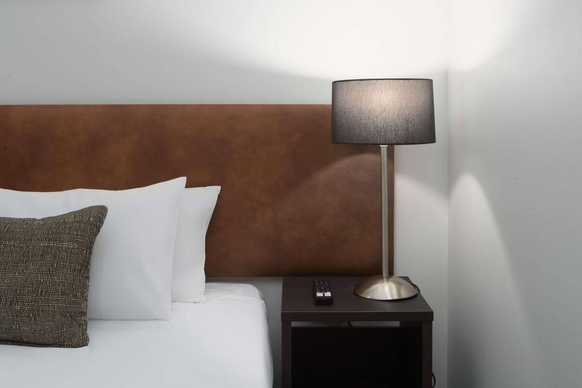 A hotel room with a bed, pillows, a nightstand, a remote control, and a lamp.