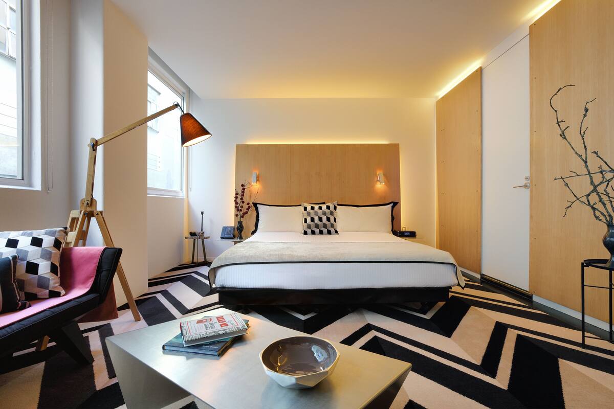 A modern hotel room with a large bed, a desk with a bowl, a lamp, and a checkered rug.