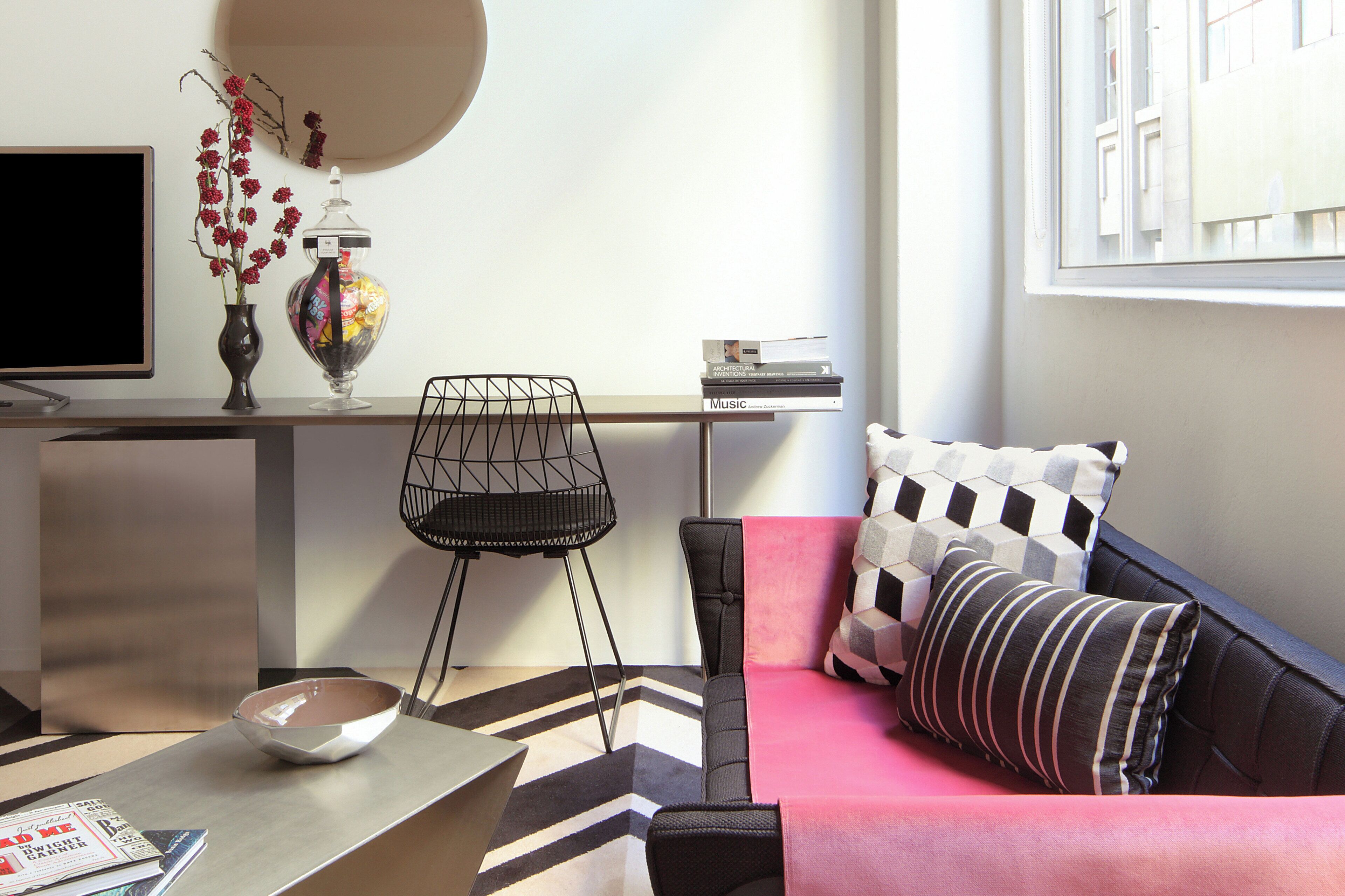 A modern living room with a flat-screen TV, a coffee table, a sofa with patterned cushions, a decorative vase with red flowers, and a stack of books on a shelf.