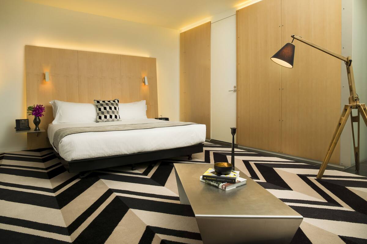 A modern hotel room with a large bed, a bedside table with a lamp, a small table with books, and a geometric patterned rug.