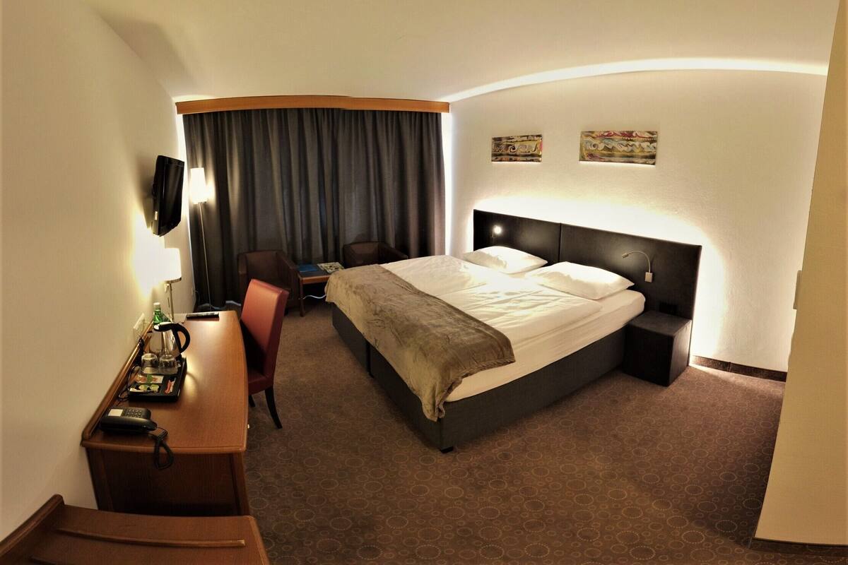 Habitación de hotel con una cama, un escritorio con silla, televisor y ventana con cortinas.