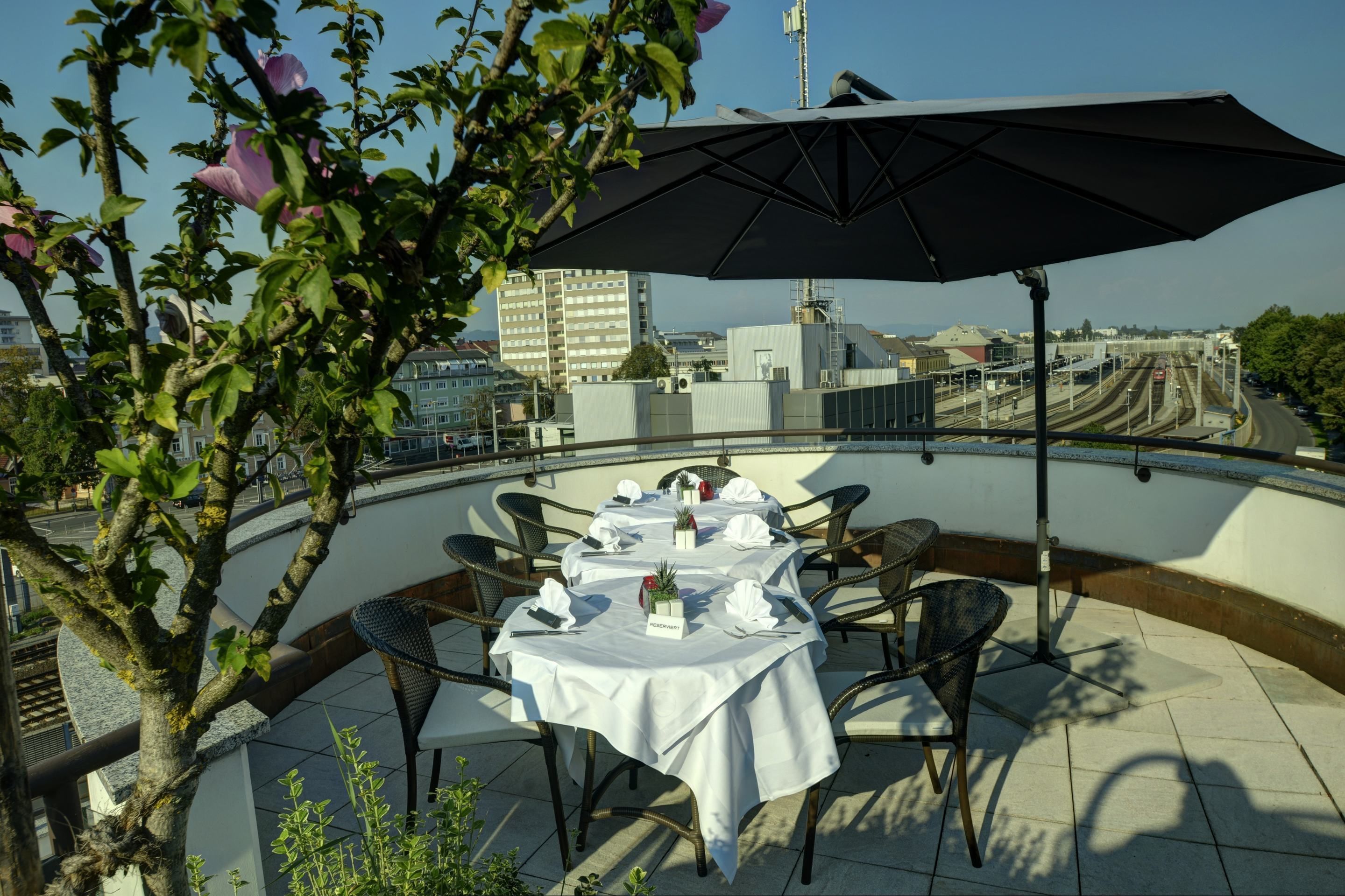 A rooftop dining area with a table set for four, surrounded by wicker chairs, under a large umbrella.