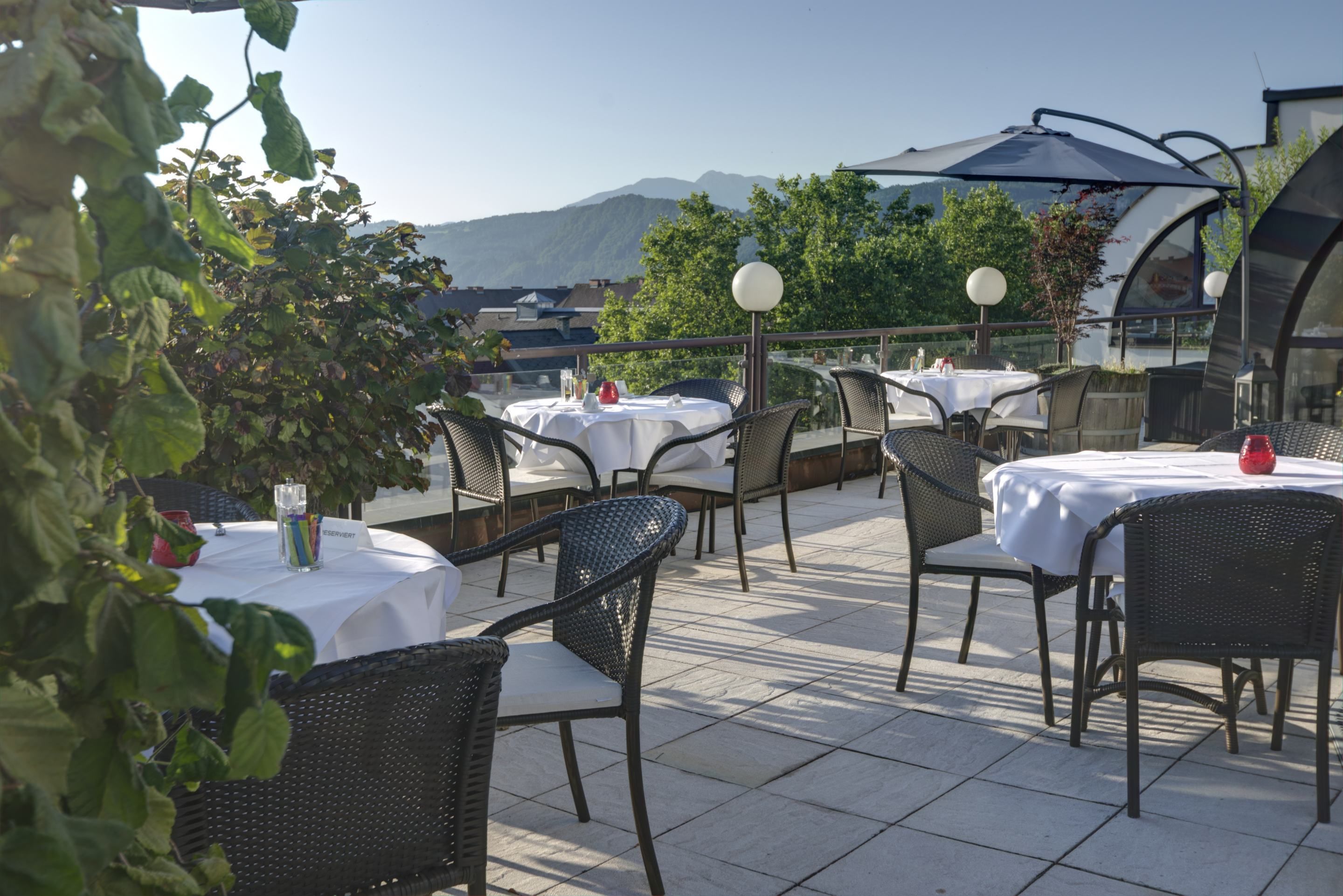 An outdoor patio with round tables and wicker chairs, set against a backdrop of mountains and trees.