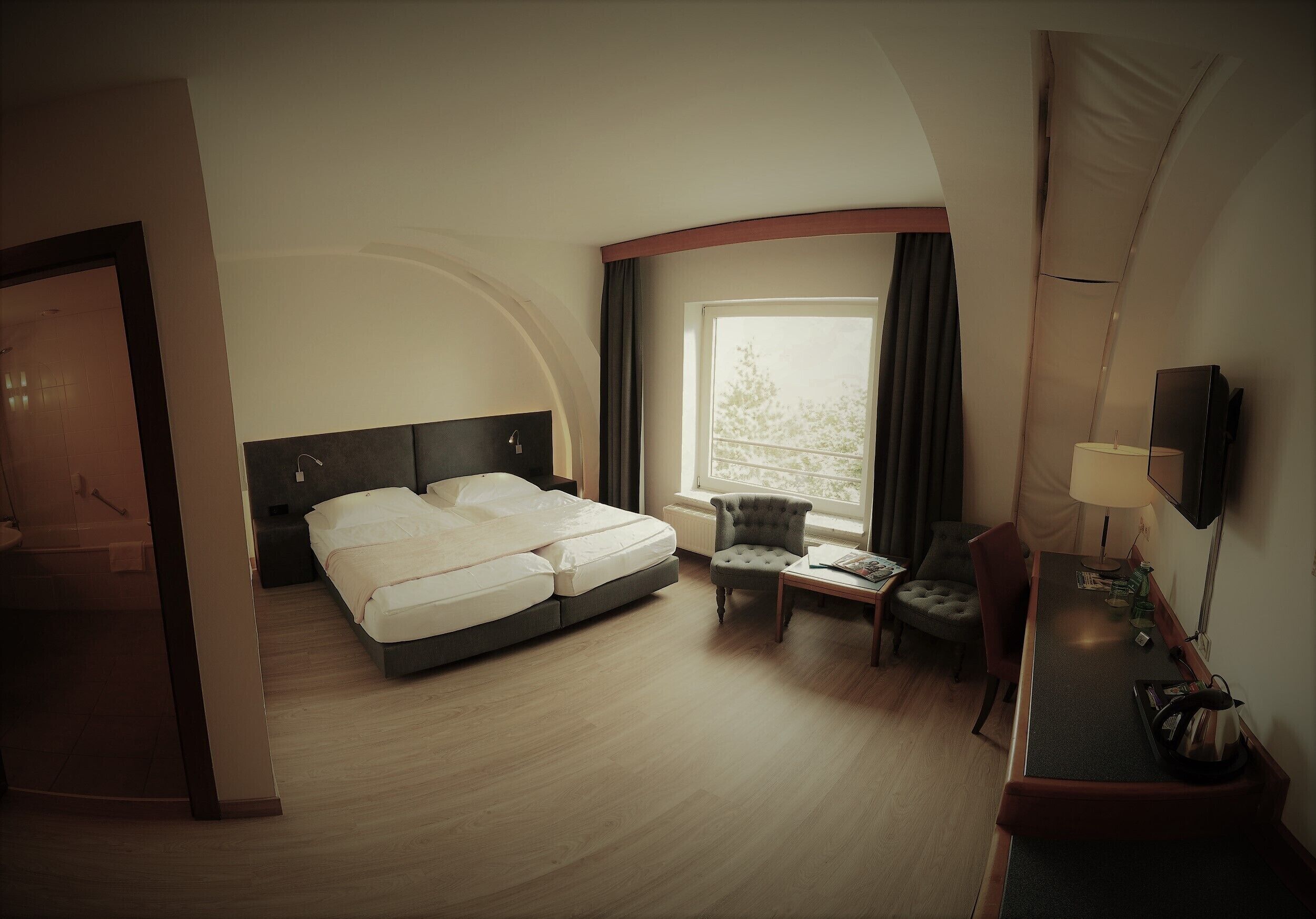 A hotel room with a bed, a small armchair, a desk with a lamp, and a TV.