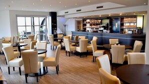 A modern restaurant with round tables and beige chairs, a bar area with shelves of bottles, and a large window providing natural light. - Skycity Hotel Atrigon (Klagenfurt am Woerthersee)