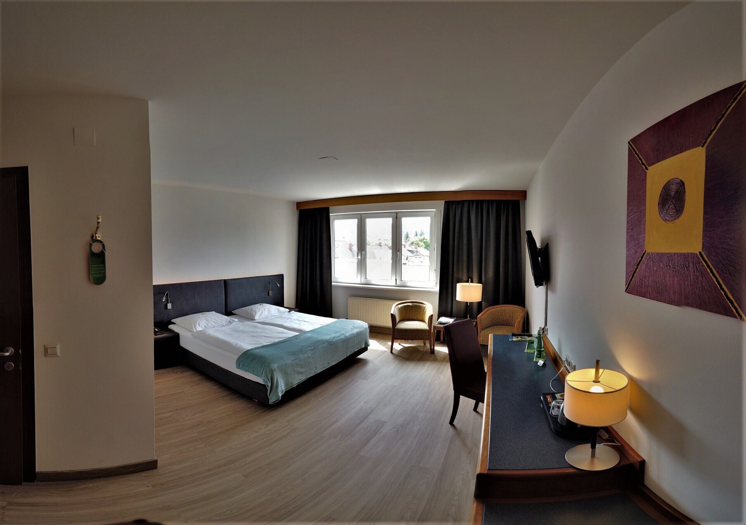 A hotel room with a bed, a desk with a lamp, a chair, and a TV mounted on the wall.