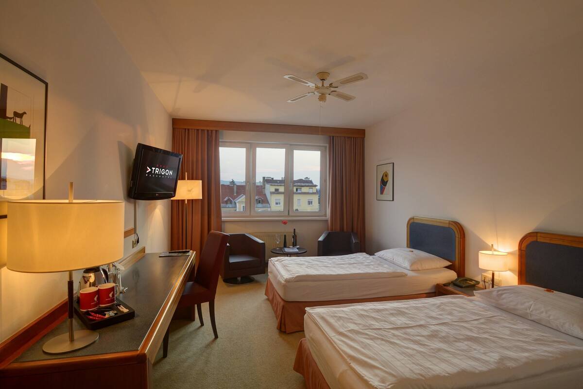 Habitación de hotel con dos camas, un escritorio con lámpara, televisor y vistas a edificios a través del ventanal.