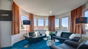 A modern living room with a curved ceiling, large windows, and a flat-screen TV mounted on the wall. - Skycity Hotel Atrigon (Klagenfurt am Woerthersee)