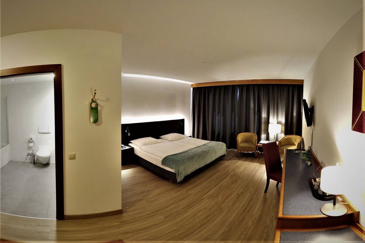 Habitación de hotel con cama, escritorio, silla, espejo y baño visible a través de una puerta abierta.