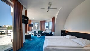 A modern hotel room with a large bed, a sofa, a TV, and a balcony with a view of the city. - Skycity Hotel Atrigon (Klagenfurt am Woerthersee)