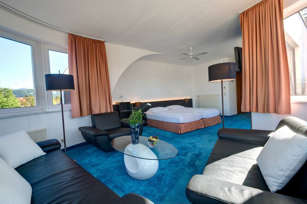 Una habitación de hotel moderna con alfombra azul, un sofá, una cama, un televisor y un ventilador de techo.