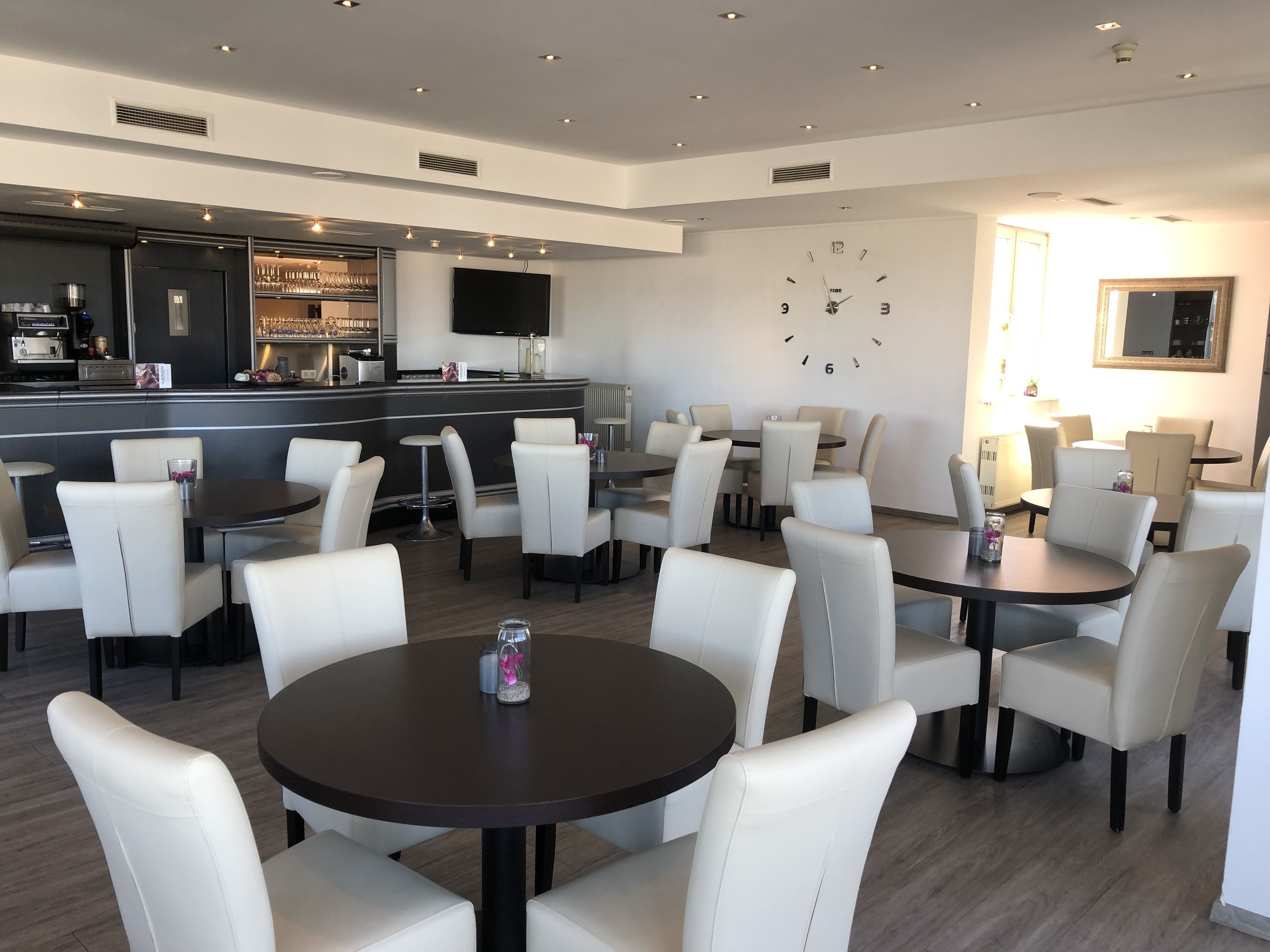 Eine moderne Hotelhalle mit runden Tischen und weißen Ledersesseln, einem Barbereich mit Fernseher sowie einer Uhr an der Wand.