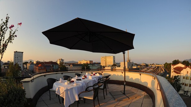A rooftop terrace with tables and chairs, a large umbrella, and a cityscape view in the background. - Skycity Hotel Atrigon (Klagenfurt am Woerthersee)