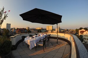 A rooftop terrace with tables and chairs, a large umbrella, and a cityscape view in the background. - Skycity Hotel Atrigon (Klagenfurt am Woerthersee)