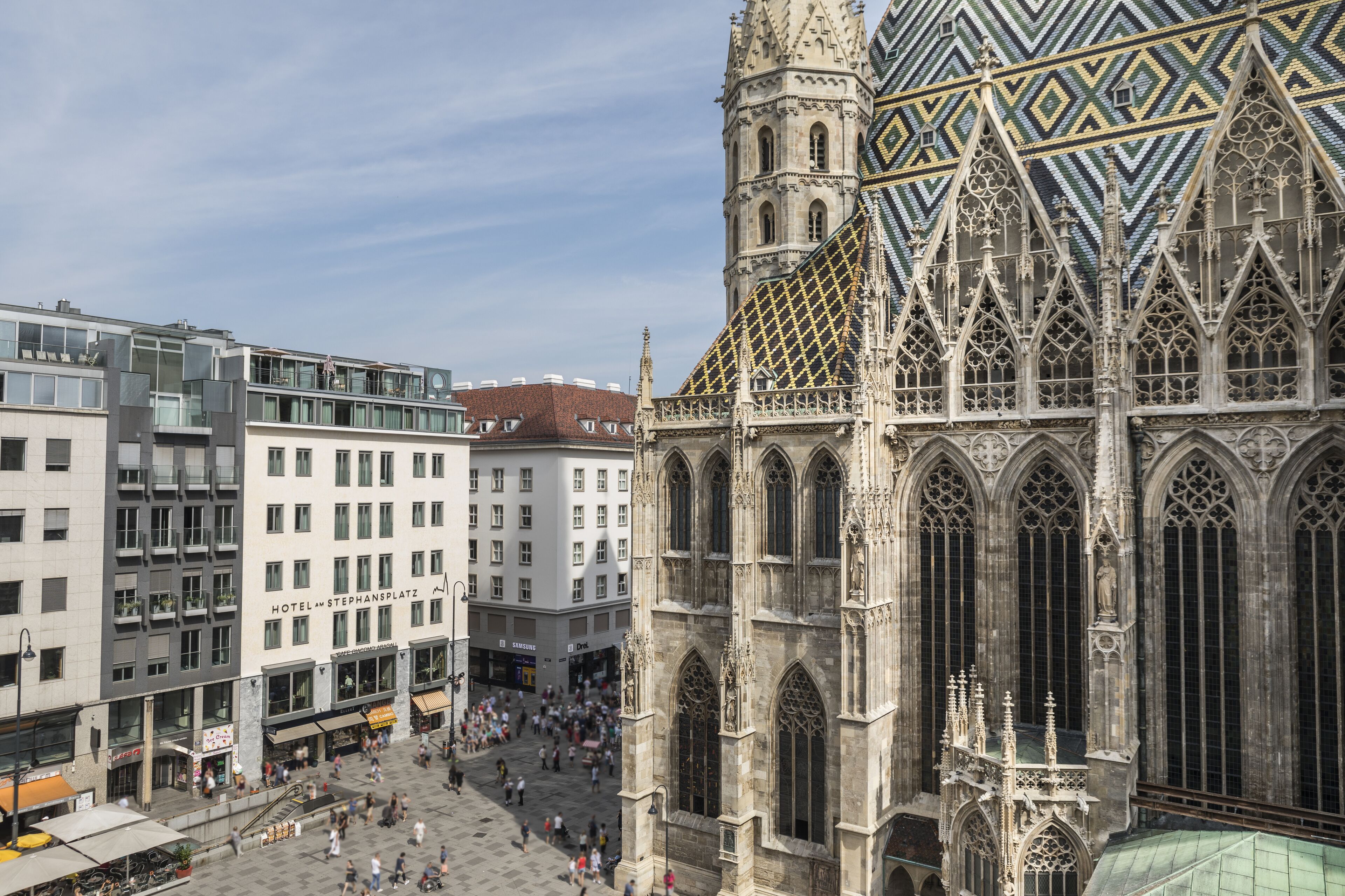 Una catedral histórica con arquitectura gótica detallada, un hotel moderno contiguo a ella, y una plaza animada con gente caminando.