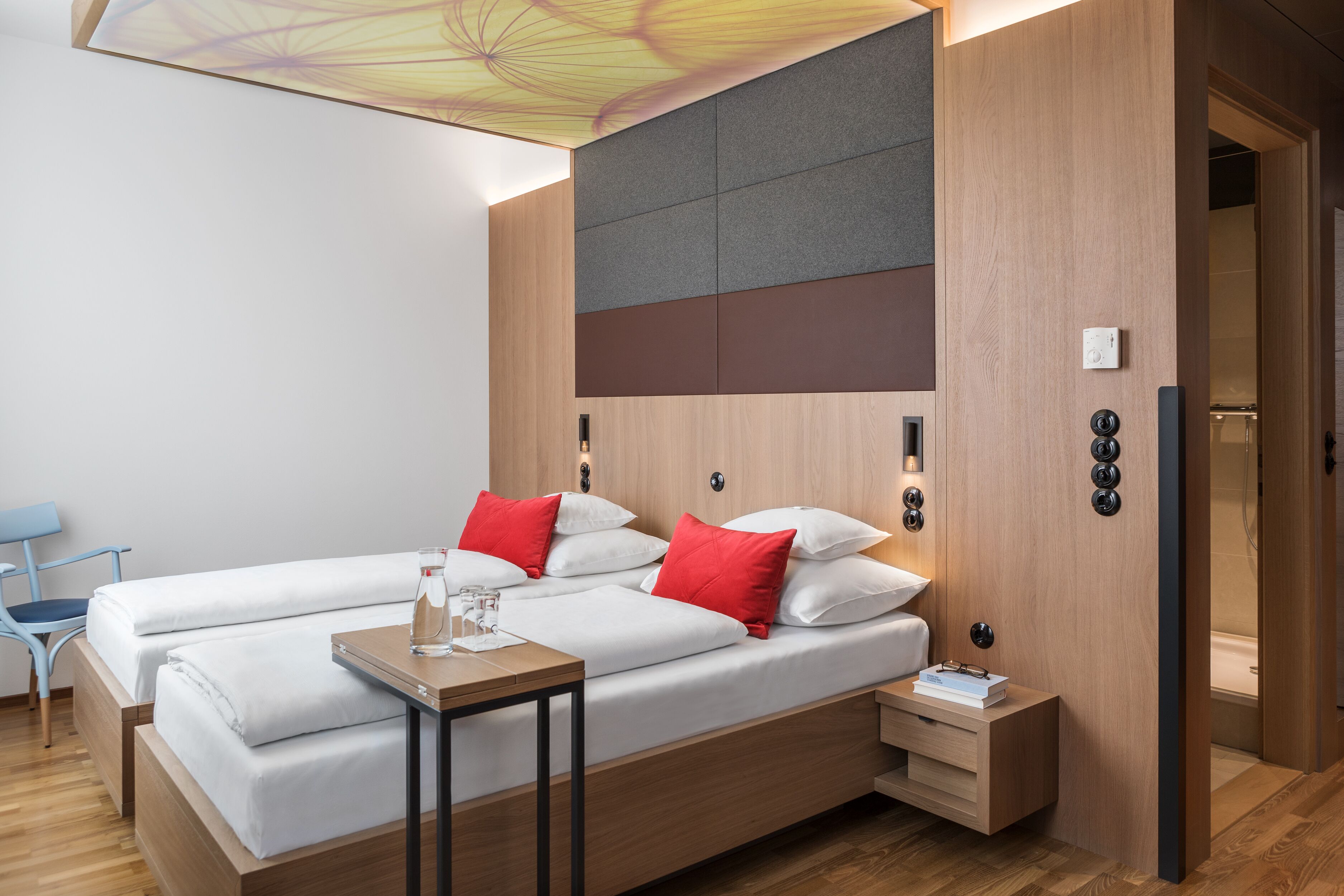 Quarto de hotel moderno com cama de madeira, duas mesas de cabeceira e luminária no teto.