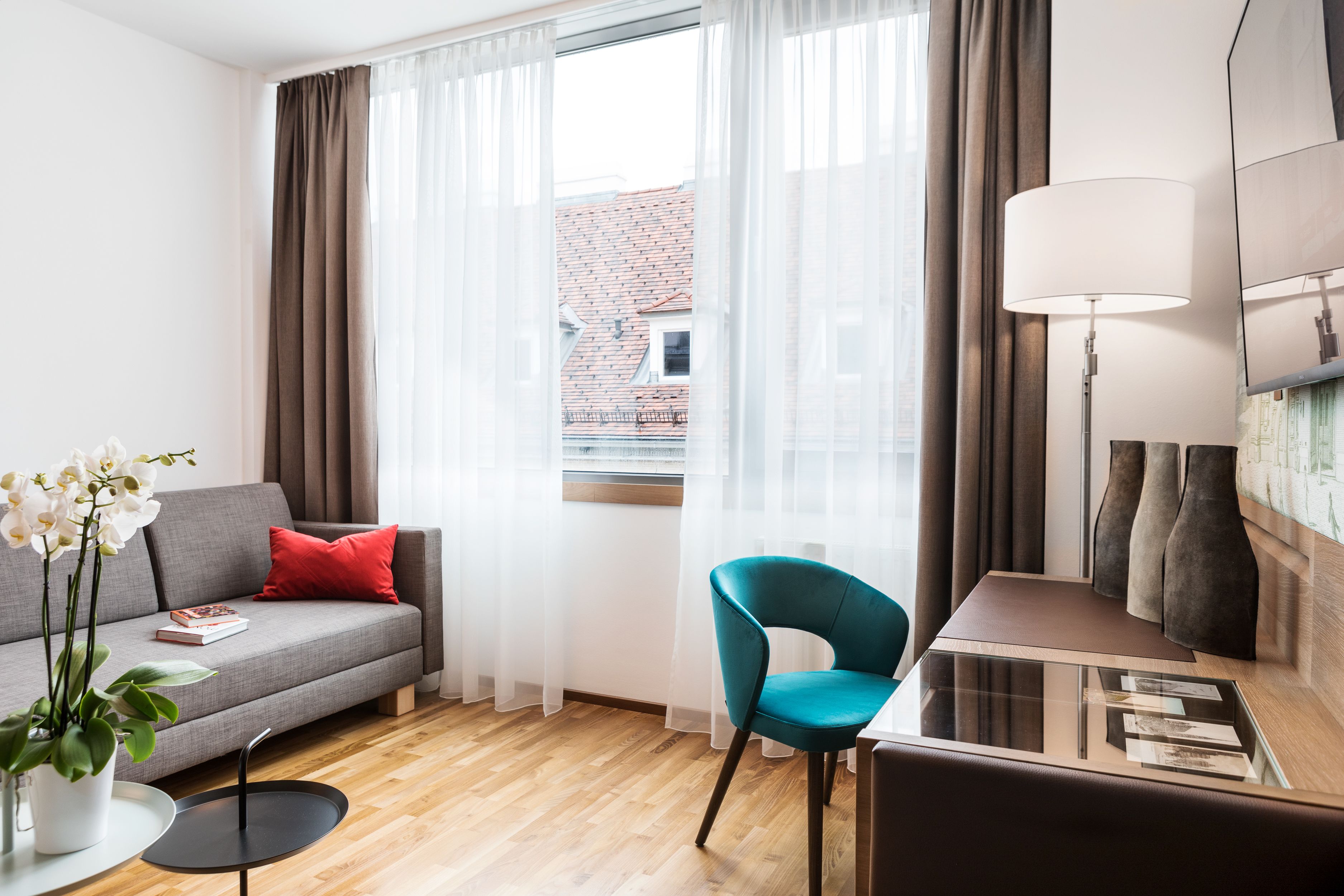 A modern living room with a grey sofa, red pillow, teal armchair, and a small table with a lamp.