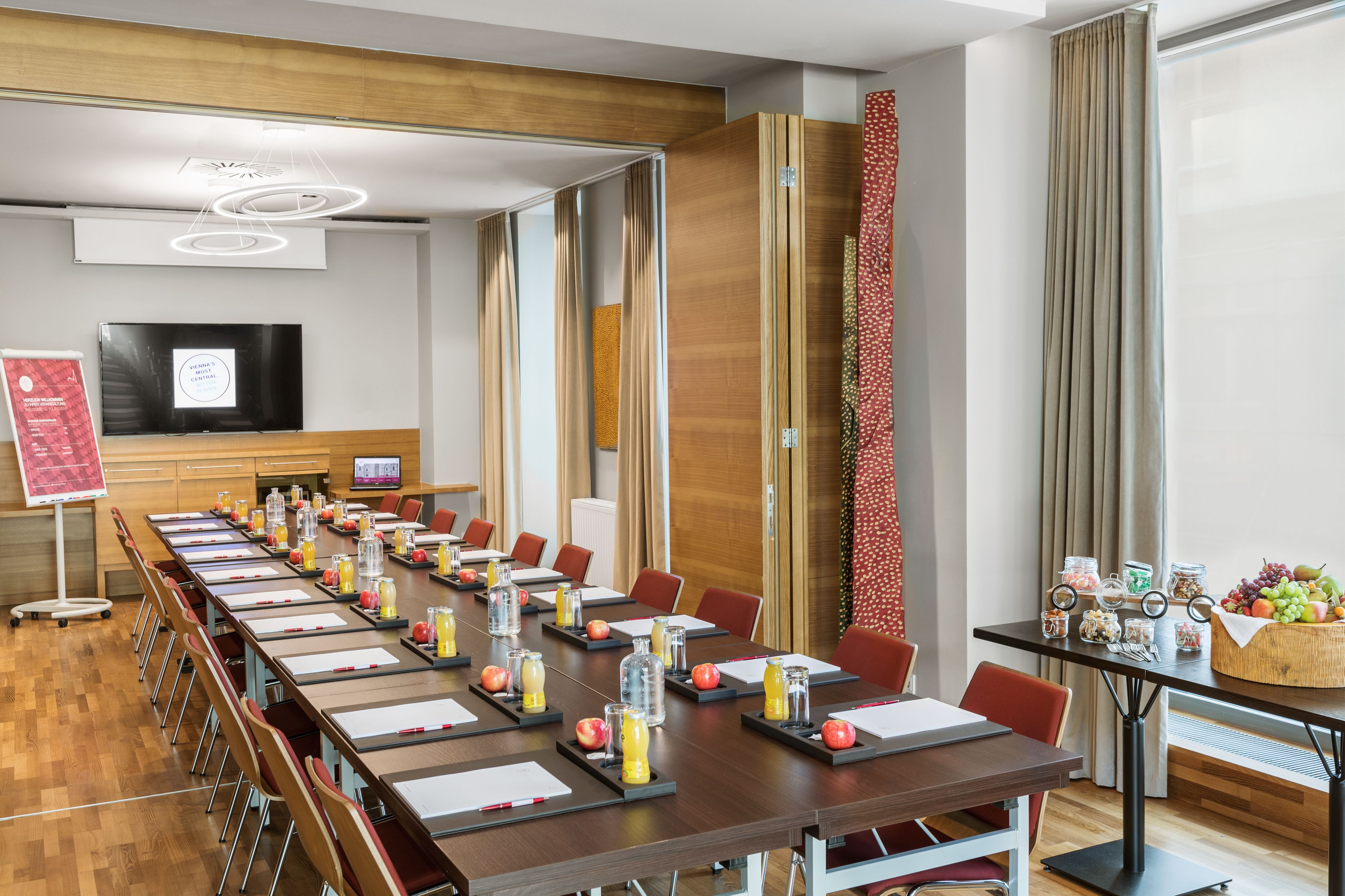 Un salón de conferencias con una larga mesa, sillas, una pantalla de proyector y una mesa de buffet con frutas y aperitivos.