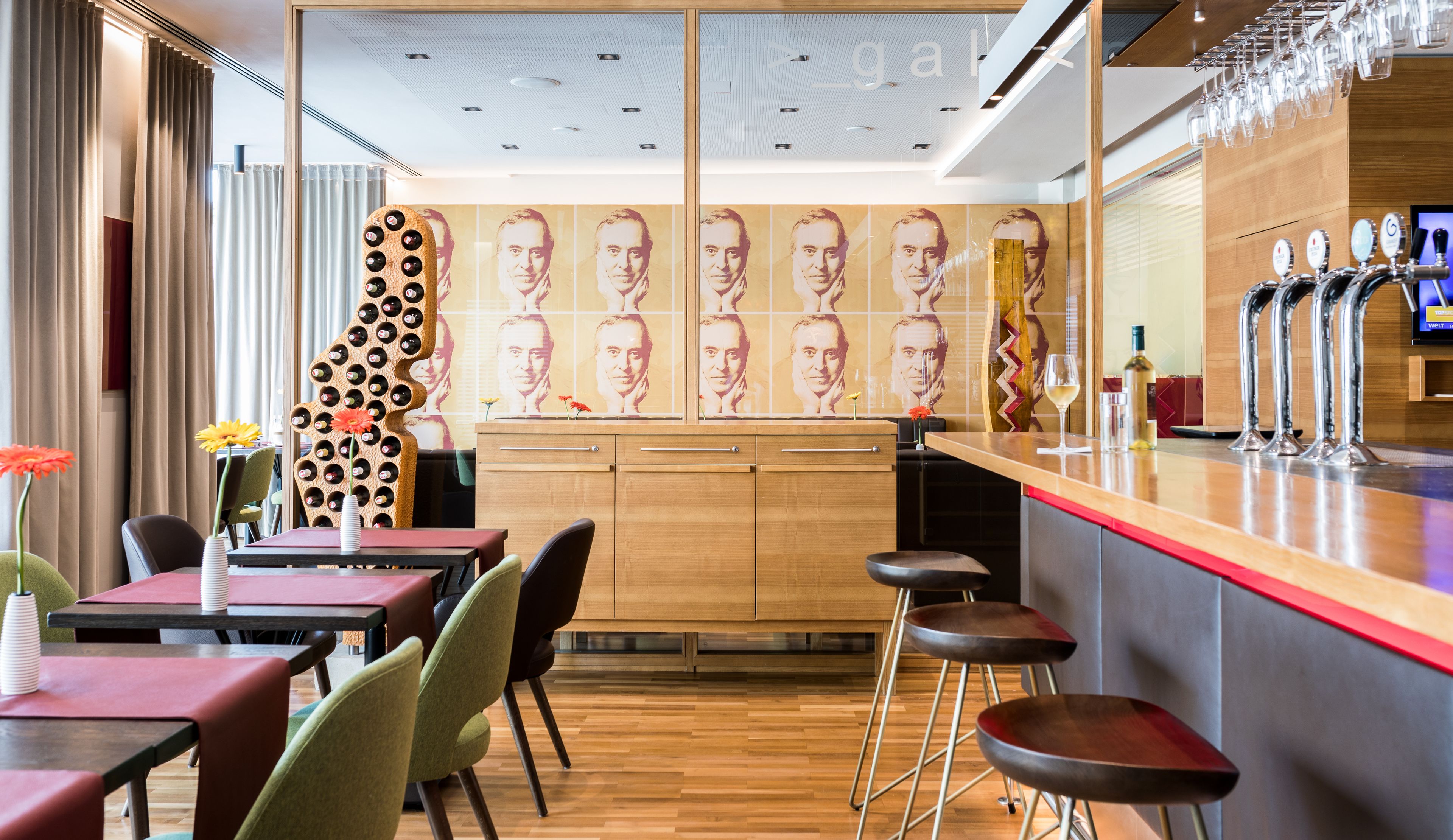 A modern bar area with a wooden counter, multiple bar stools, and a wall with repeated portraits.