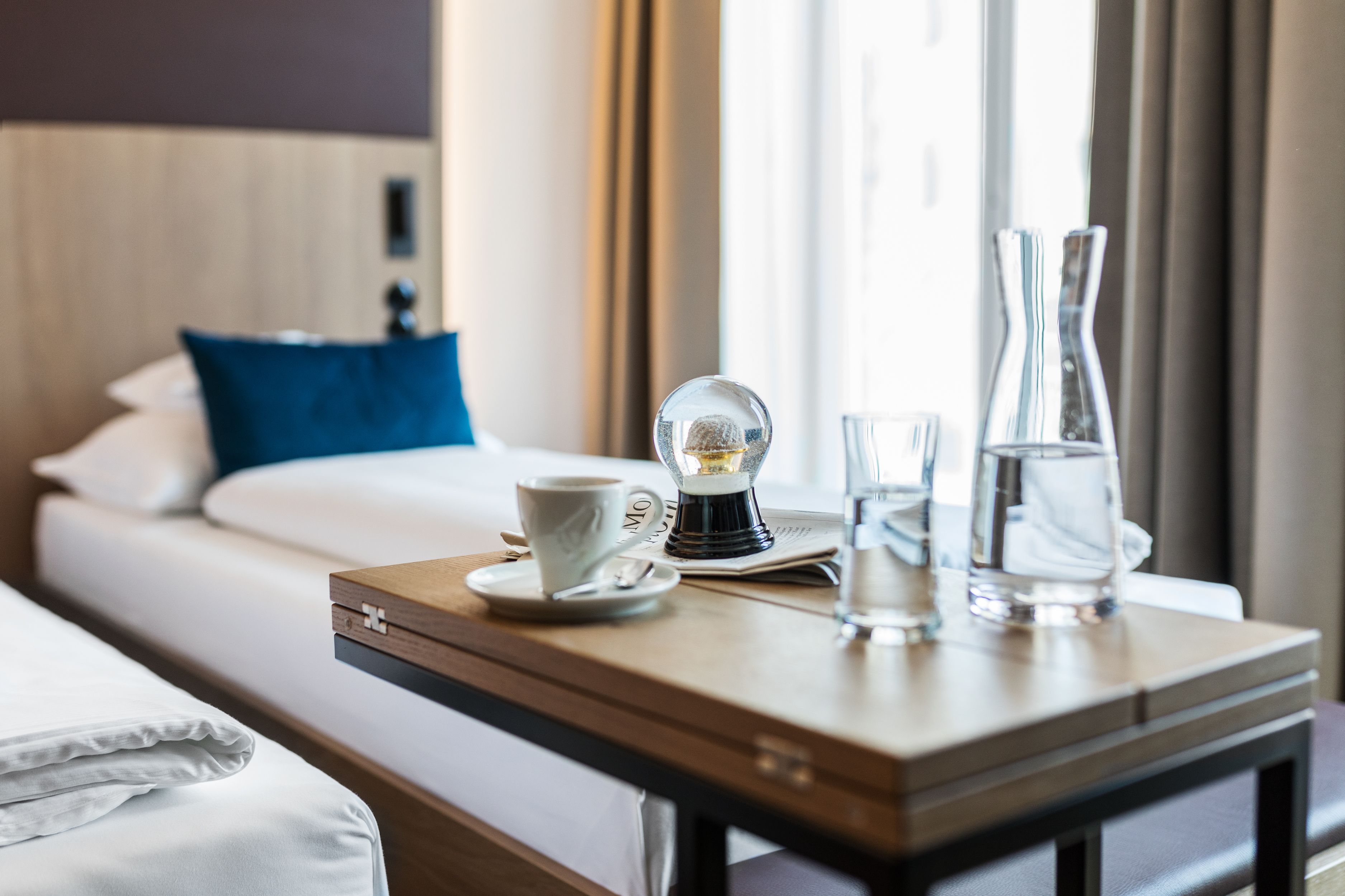 A neatly made bed with a blue pillow, a coffee maker, a cup, and glasses on a wooden table in a hotel room.