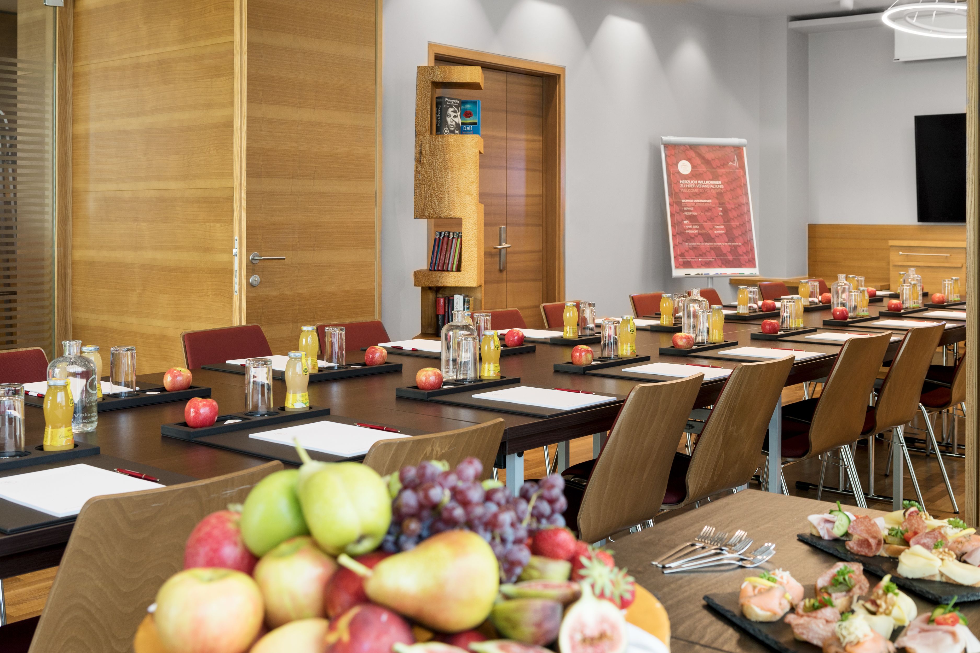 Un salón de conferencias con una larga mesa, sillas, una estantería y una pantalla para presentaciones.