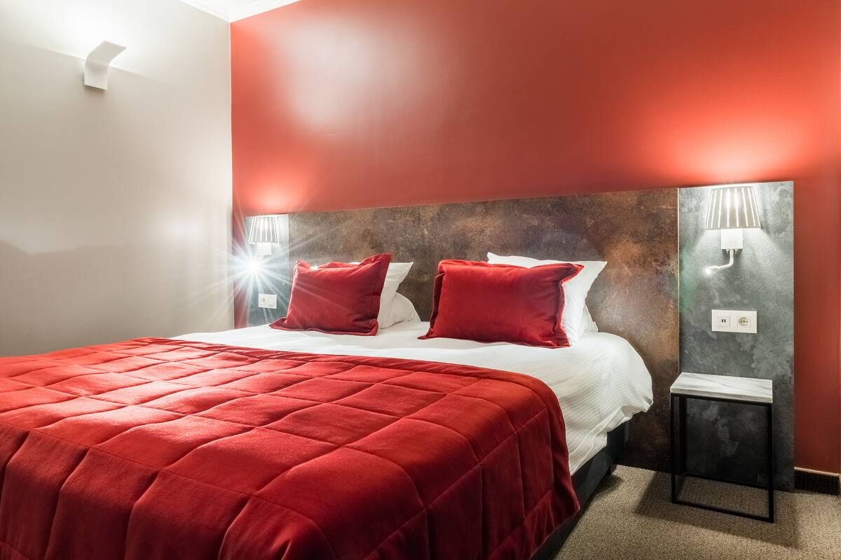 A hotel room with a red bedspread, two red pillows, a white bed, and a nightstand with a lamp.