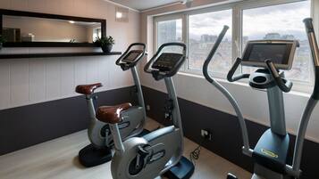 A gym with two stationary bikes and a treadmill, a large window with a city view, and a mirror on the wall.