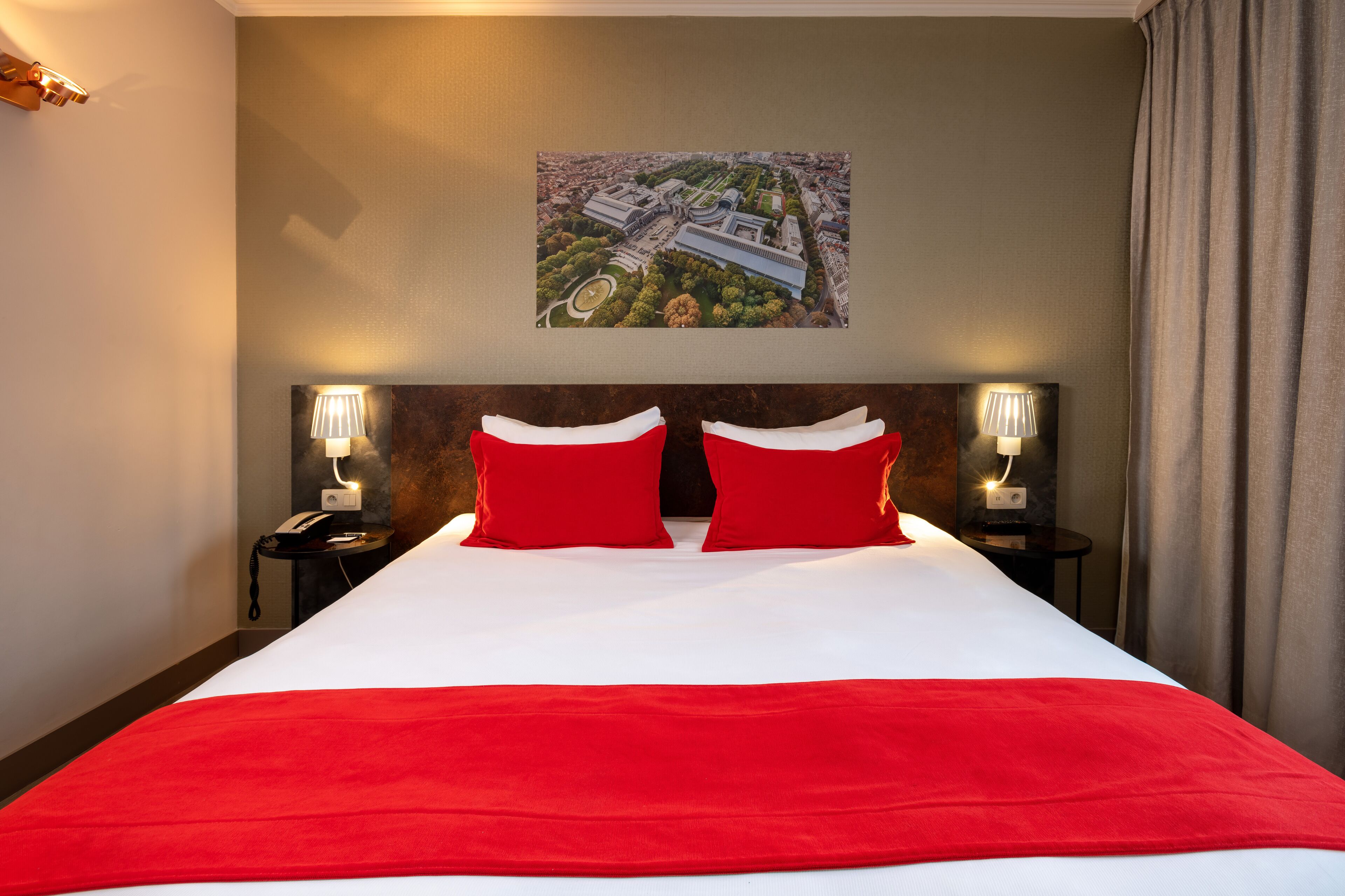 Habitación de hotel con una cama grande, almohadas rojas, cabecera con fotografía de un paisaje urbano, lámparas de noche y vista a la ciudad por la ventana.