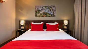 Habitación de hotel con una cama grande, almohadas rojas, cabecera con fotografía de un paisaje urbano, lámparas de noche y vista a la ciudad por la ventana.