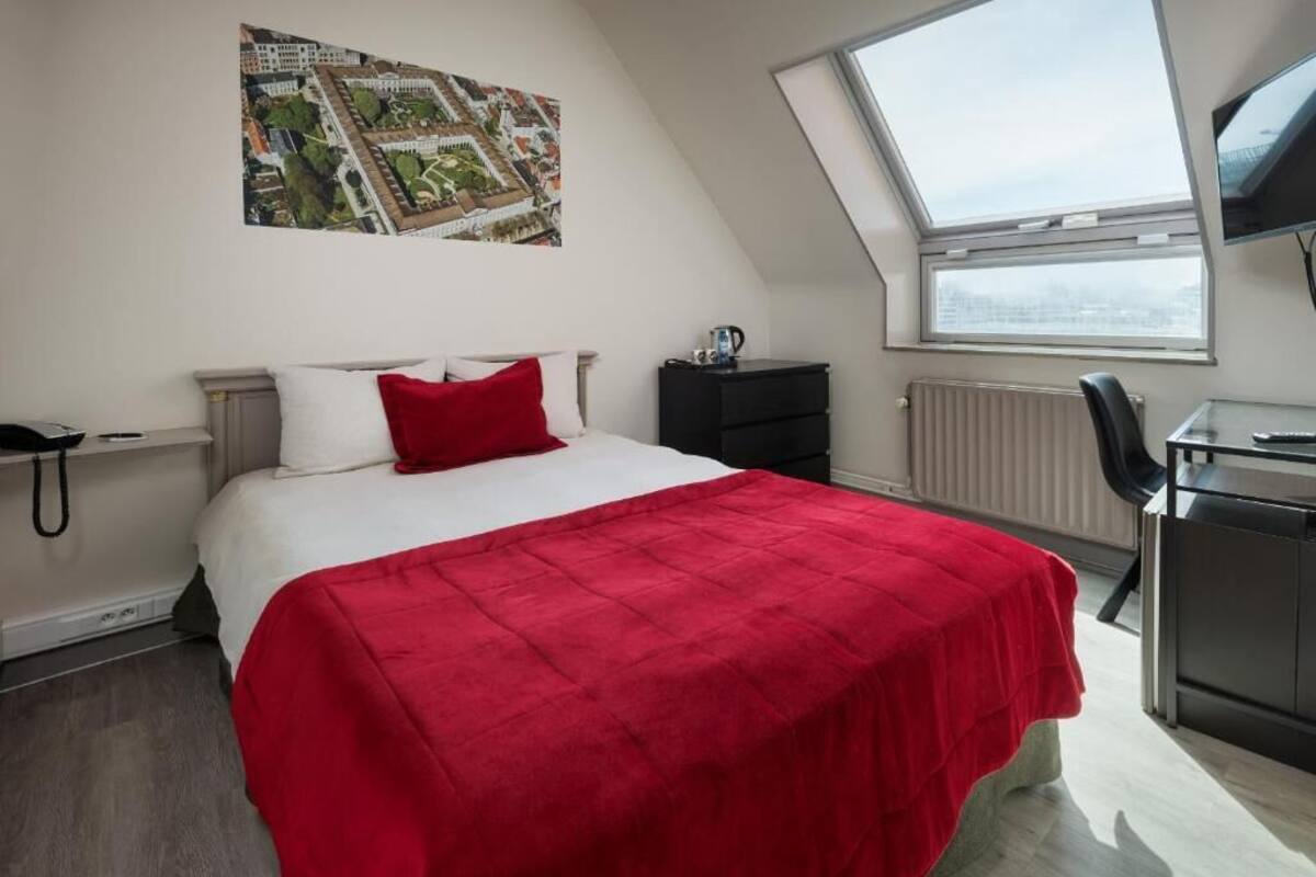 A bedroom with a bed, a red blanket, a desk with a chair, a radiator, and a window with a view of the city.