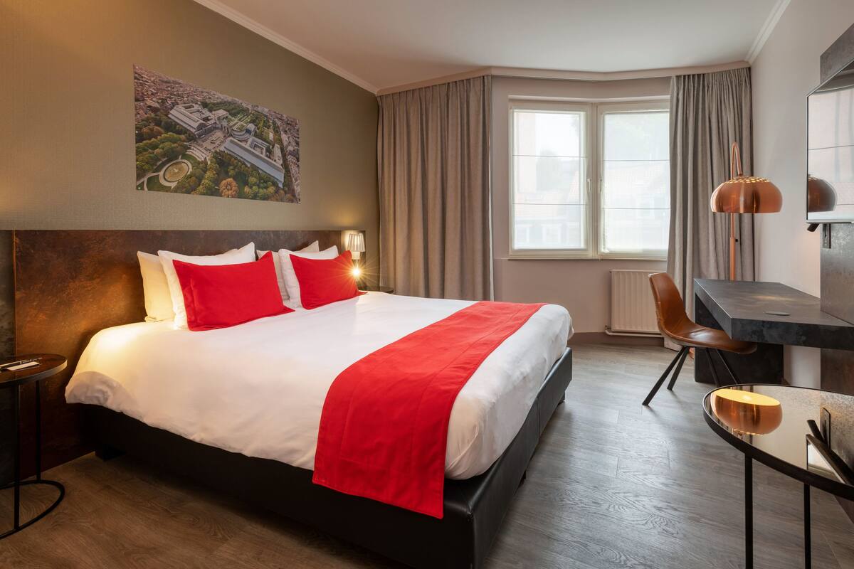 A hotel room with a large bed, a desk, a chair, a lamp, and a window with curtains.