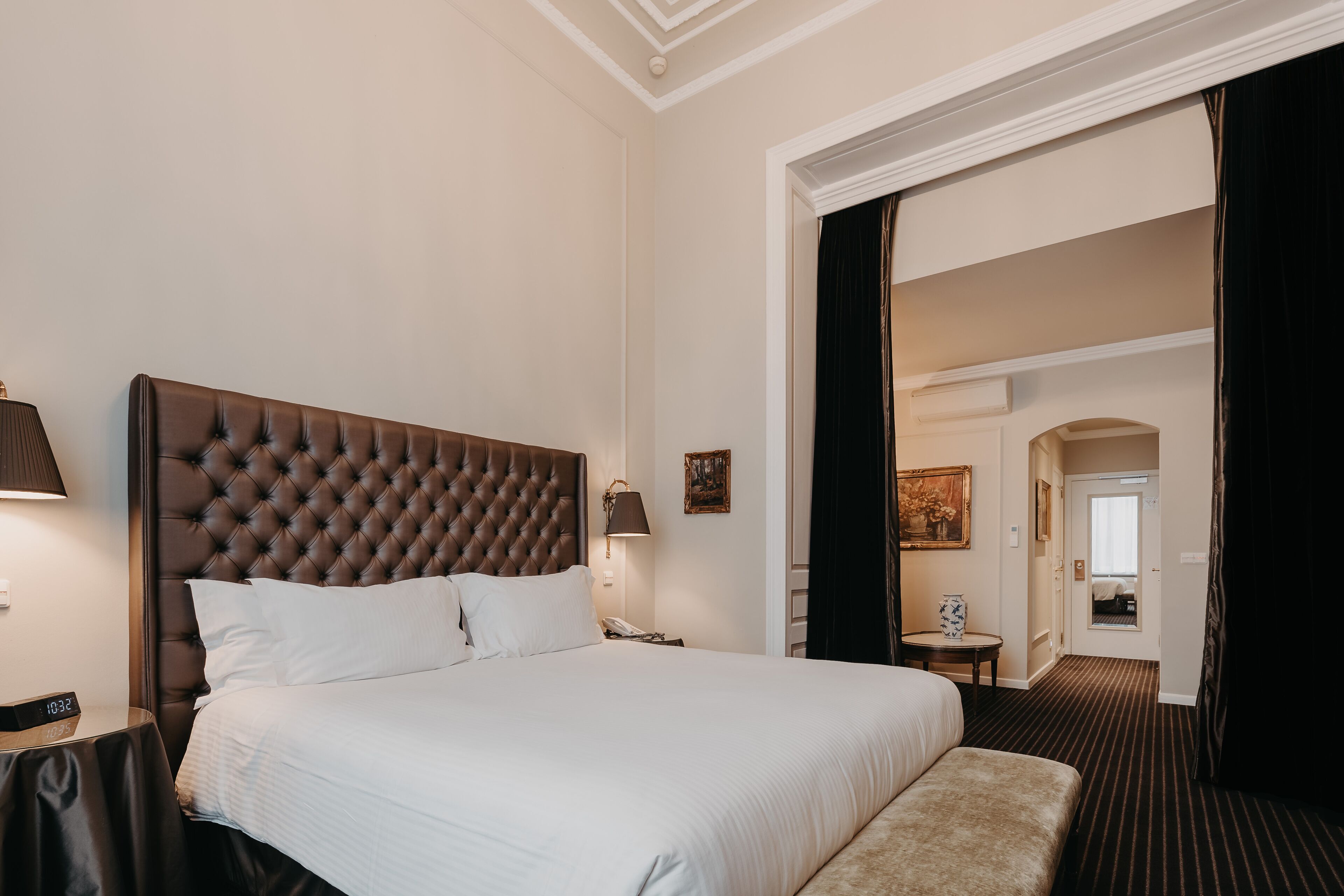 Habitación de hotel con una cama grande, una mesita de noche con lámpara, una pequeña mesa con un jarrón y una puerta que da a otra habitación.