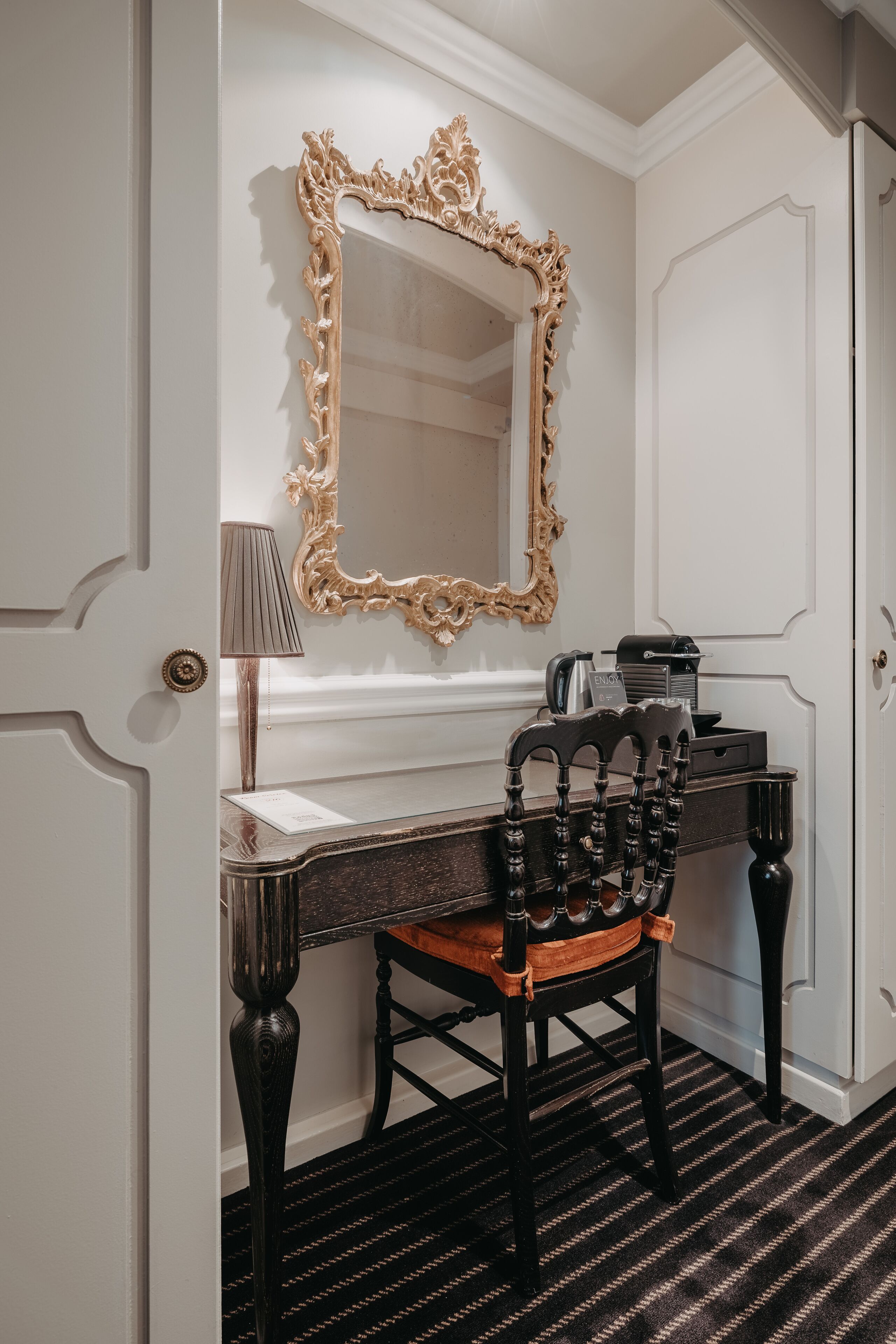 Une pièce comprenant un bureau, une chaise, une lampe et un miroir doté d’un cadre orné.