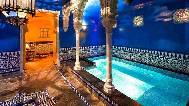 Un espace piscine intérieur luxueux, agrémenté de colonnes décoratives, d’un sol à motifs et offrant une vue sur une autre pièce.