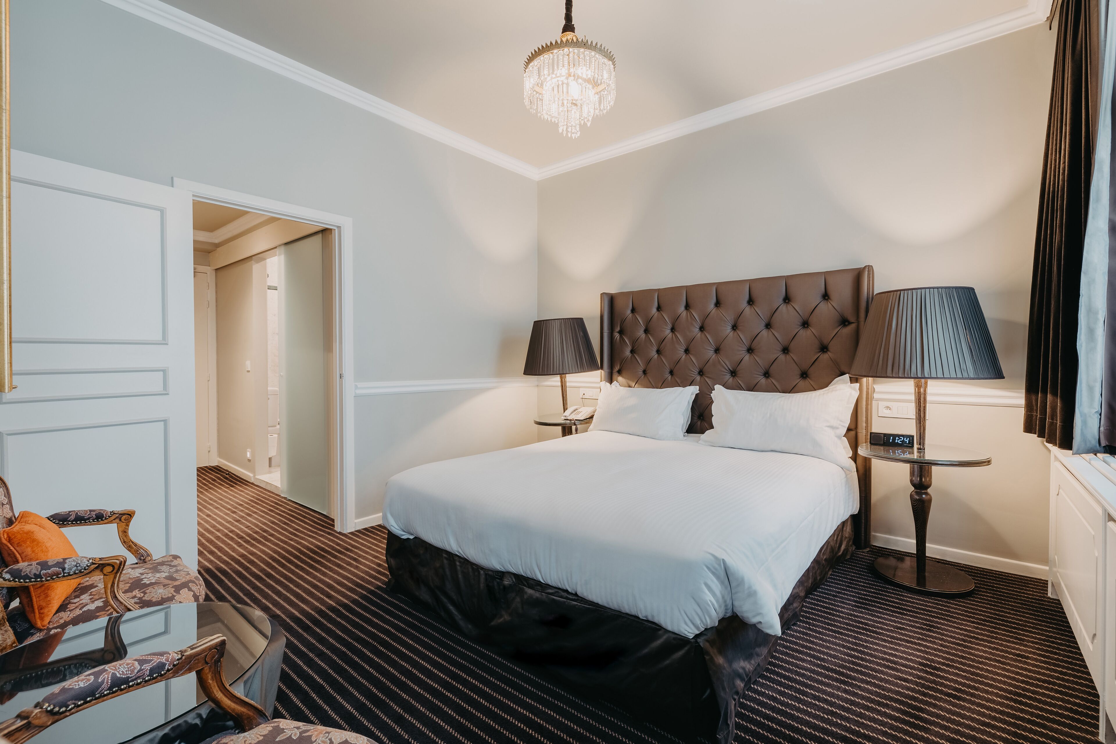 A hotel room with a large bed, two bedside lamps, a chandelier, a small table, and a chair with a cushion.