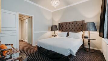 A hotel room with a large bed, two bedside lamps, a chandelier, a small table, and a chair with a cushion.