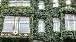 La façade d’un hôtel est recouverte de lierre, et une horloge ainsi que le nom « HÔTEL MANOS » sont visibles au-dessus de l’entrée.