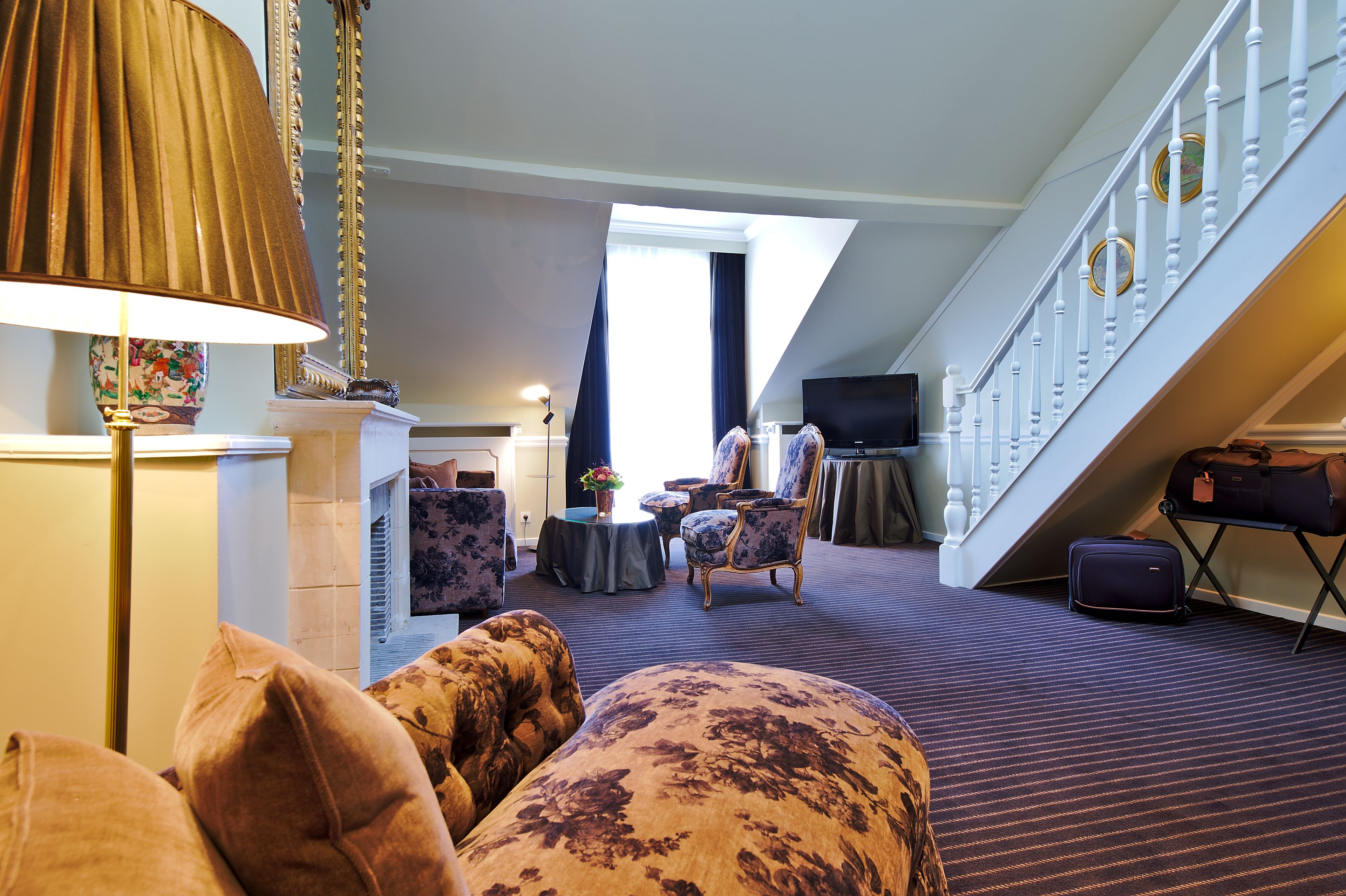 A hotel room with a living area featuring a sofa, armchairs, a lamp, and a television. A staircase with a railing and a suitcase is visible in the background.