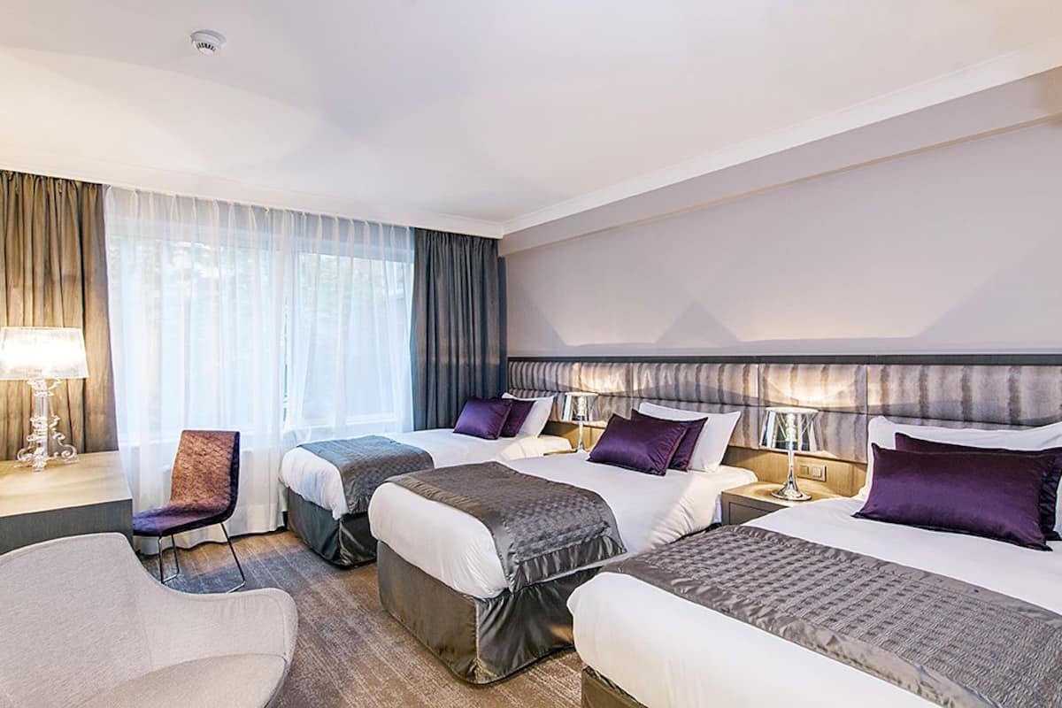 Habitación de hotel con tres camas, un escritorio, una silla y un ventanal grande con cortinas.