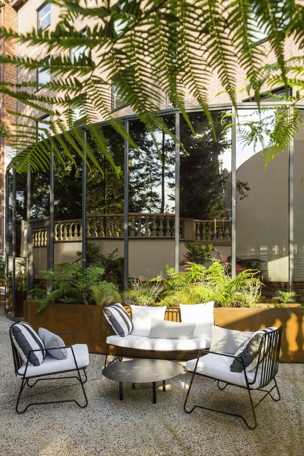 An outdoor seating area with a white sofa and two chairs, surrounded by greenery and a glass enclosure reflecting trees.