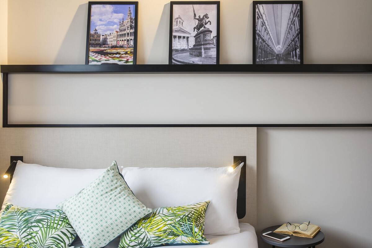 Ein Hotelzimmer mit einem Bett, Kissen, einem Nachttisch mit Buch und Gläsern sowie drei gerahmten Bildern an der Wand.