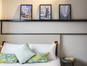A hotel room with a bed, pillows, a bedside table with a book and glasses, and three framed pictures on the wall.