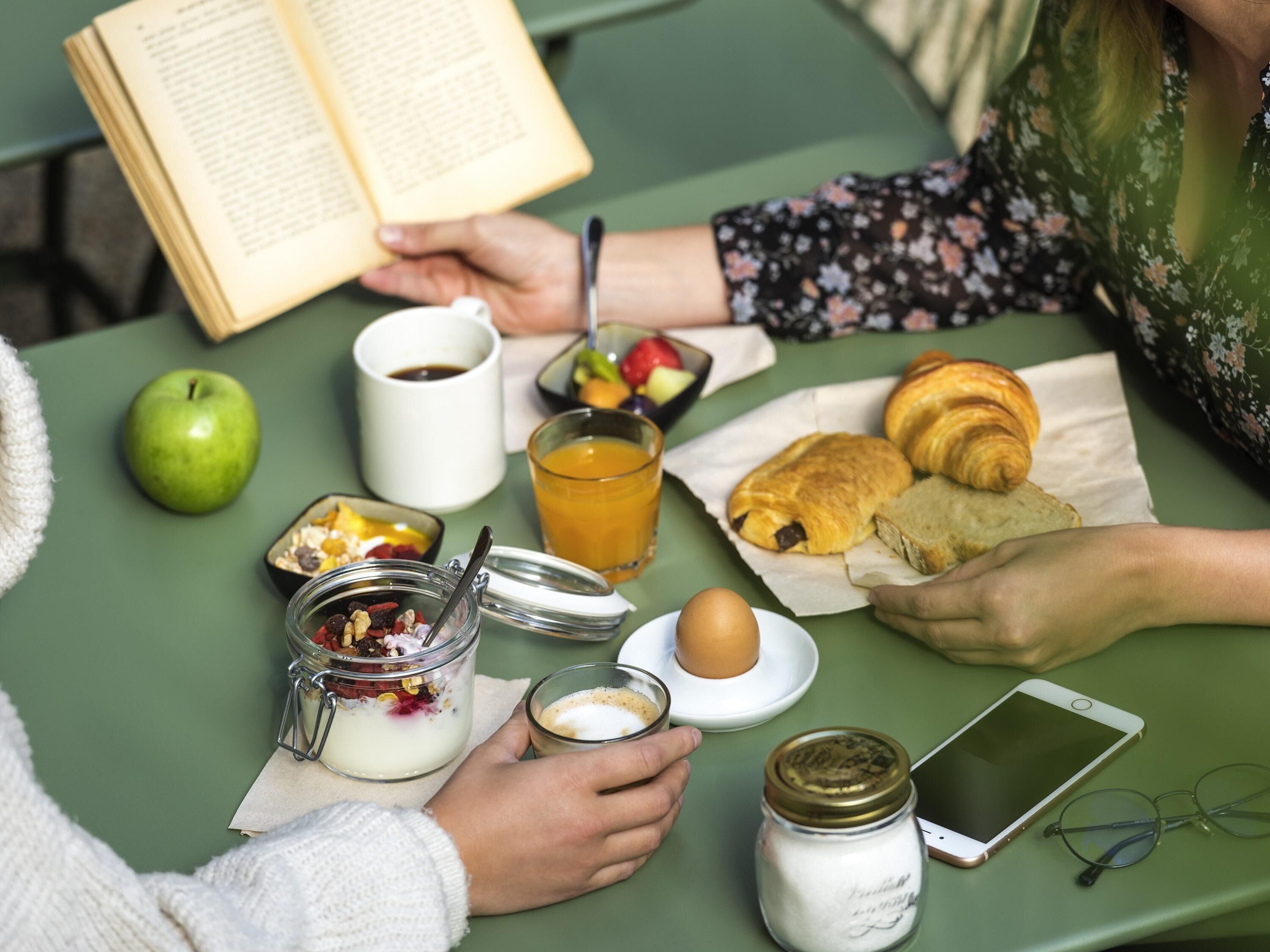 A table with a book, a glass of orange juice, a croissant, a sandwich, an apple, a jar of yogurt, a jar of sugar, a smartphone, and a pair of glasses.