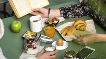 A table with a book, a glass of orange juice, a croissant, a sandwich, an apple, a jar of yogurt, a jar of sugar, a smartphone, and a pair of glasses.