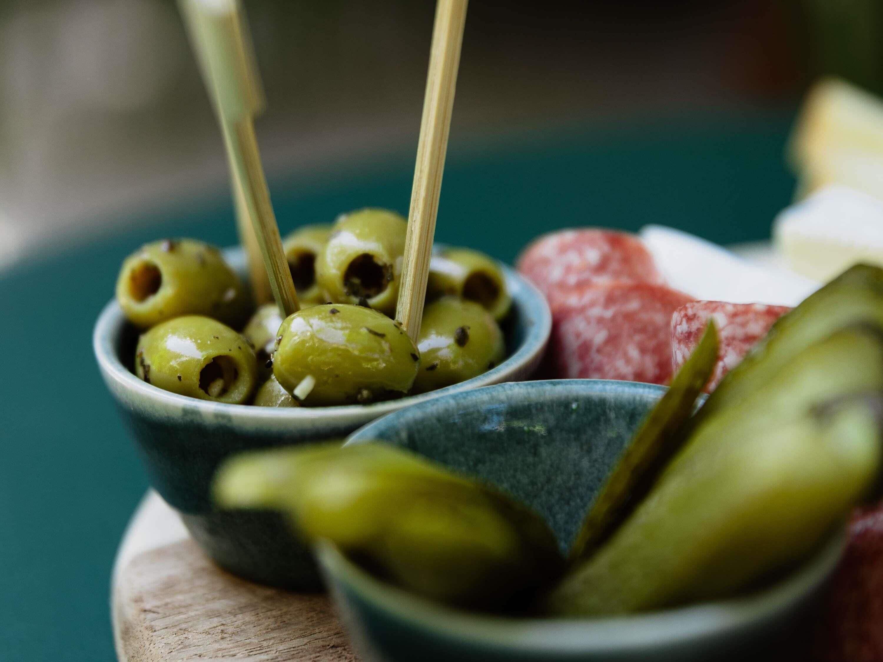 Un tazón de aceitunas con una cuchara de madera, un tazón de pepinillos y un trozo de carne sobre una tabla de madera.