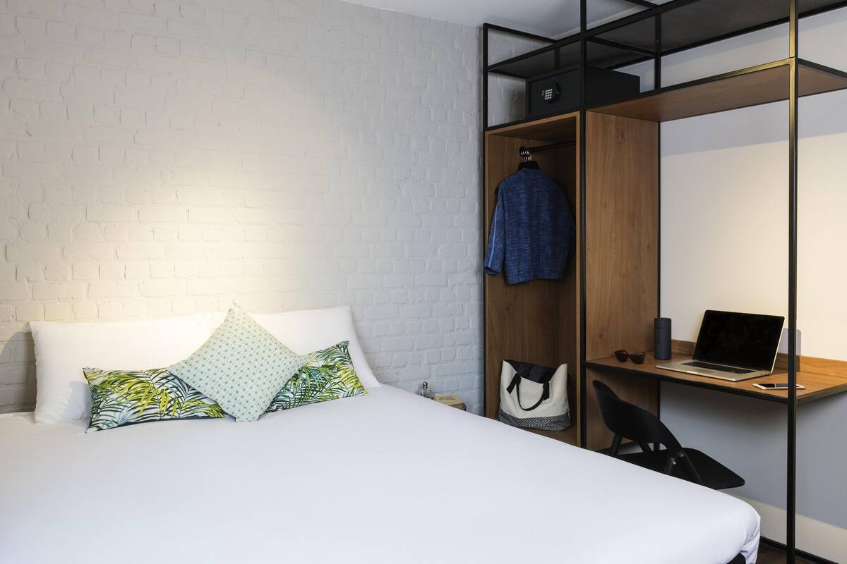 Ein modernes Hotelzimmer mit weißer Ziegelwand, einem Bett mit gemusterten Kissen, einem Holztisch mit einem Laptop und einem an der Wand befestigten Kleiderhaken.