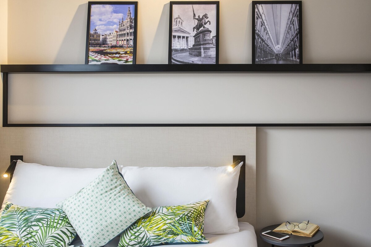 A hotel room with a bed, pillows, a bedside table with a book and glasses, and three framed pictures on the wall.