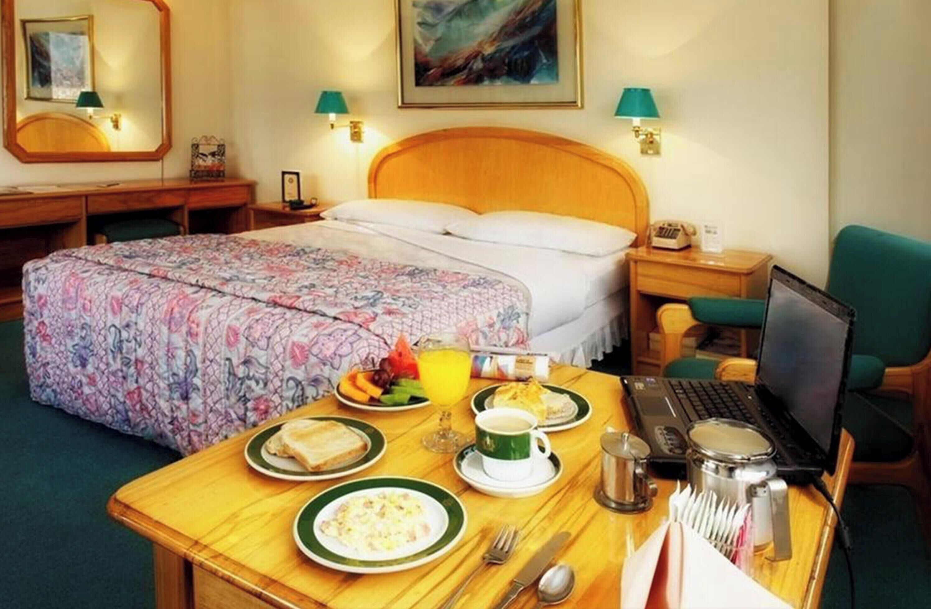 A hotel room with a bed, a desk with breakfast, a laptop, and a green chair.