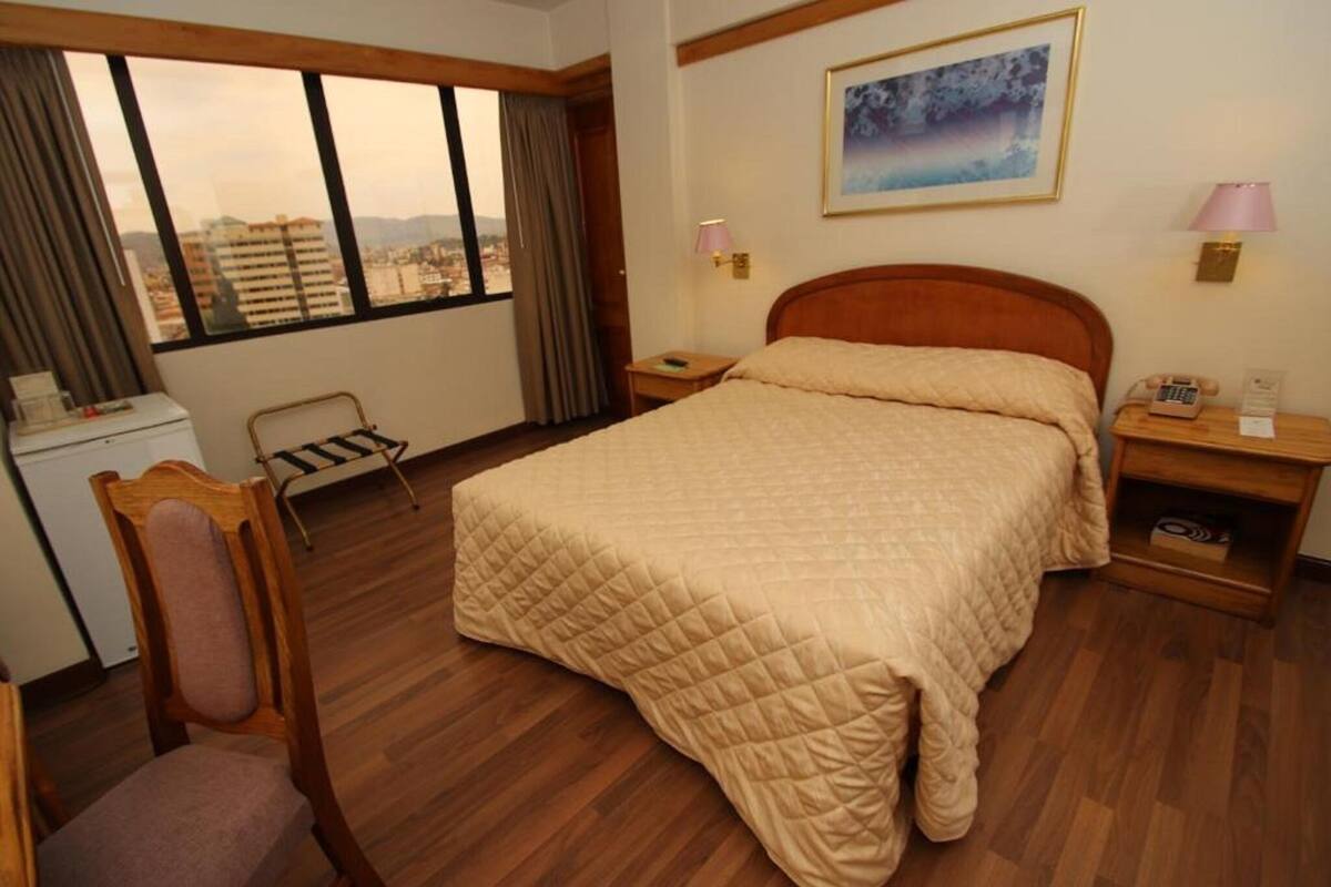 Habitación de hotel con cama, escritorio, silla, ventana con vistas y dos lámparas de noche.