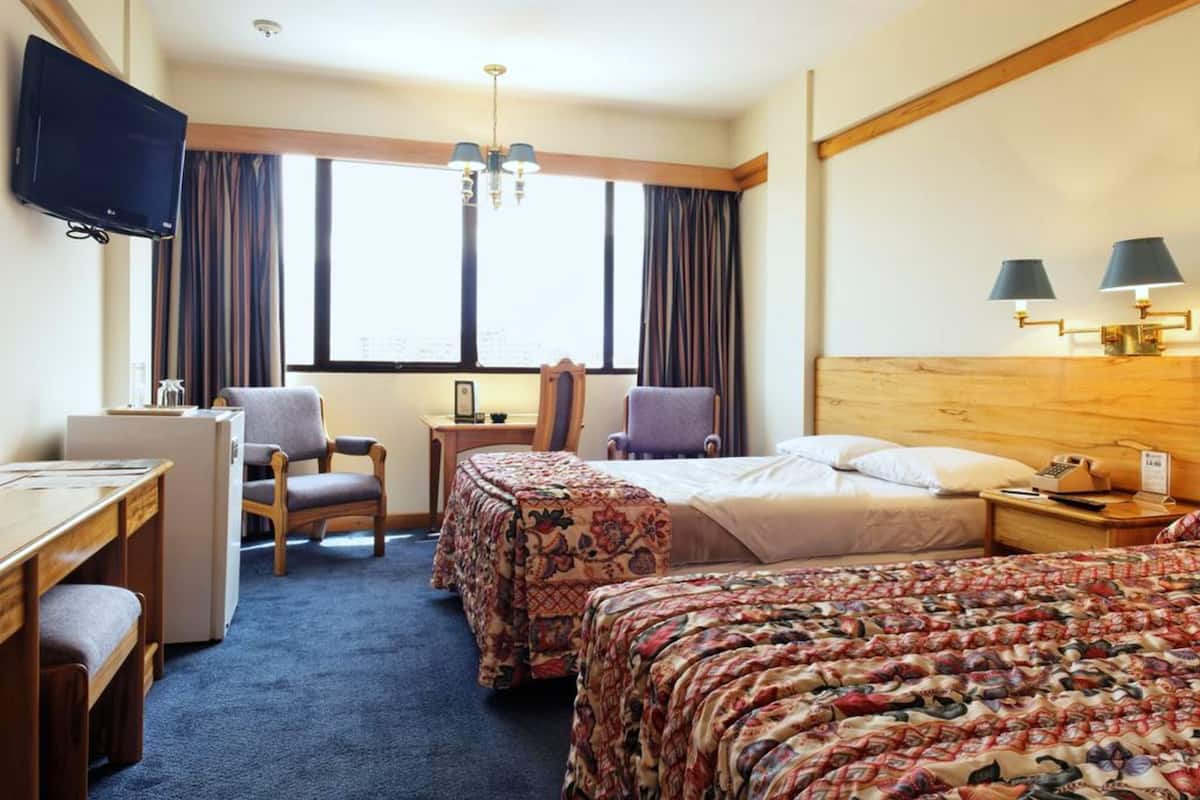 A hotel room with a desk, two chairs, a bed with floral bedding, a television, and a window with curtains.